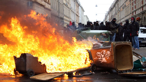 Symboldbild für Linksextremismus. Zu sehen sind vermummte Personen im Hintergrund und umgestoßene und teilweise brennende Mülltonnen im Vordergrund.