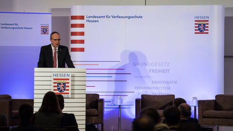 Zu sehen ist der hessische Innenminister Peter Beuth bei seiner Begrüßungsrede auf dem 23. Herbstgespräch. Beuth steht hinter einem Rednerpult. Hinter ihm sind ein Roll-Up und eine Medienwand jeweils mit der Aufschrift "Landesamt für Verfassungsschutz Hessen" zu sehen.