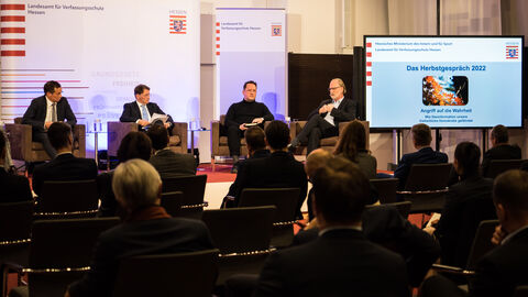 Zu sehen ist ein Blick auf das Podium des Herbstgespräches mit den Diskussionsteilnehmern Dr. Andreas Fahrner (BND), Florian Flade (Journalist), Tom Buschardt (Kommunikationstrainer) und dem Moderator Thomas Kreutzmann (Journalist). Am rechten Bildrand ist zudem ein Monitor zu sehen auf dem der Titel des Herbstgesprächs "Angriff auf die Wahrheit - Wie Desinformation unsere freiheitliche Demokratie" gezeigt wird.