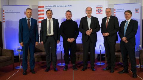Zu sehen ist ein Gruppenbild mit den Teilnehmern der Podiumsdiskussion, Thomas Kreutzmann, Florian Flade, Dr. Andreas Fahrner und Tom Buschardt, sowie dem kommissarischen Behödernleiter des LfV Hessen und dem hessischen Innenminister Peter Beuth. Nicht im Bild zu sehen ist die Podiumsteilnehmerin Julia Smirnova. 