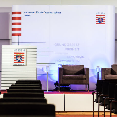 Das Herbstgespräch des LfV Hessen - Symbolbild Zu sehen ist ein bestuhlter Konferenzsaal. Man blickt auf eine Bühne auf der ein Redenerpult und mehrere Sessel zu sehen sind. Dahinter Roll-Ups und eine Medienwand jeweils mit der Aufschrift "Landesamt für Verfassungsschutz" Hessen.
